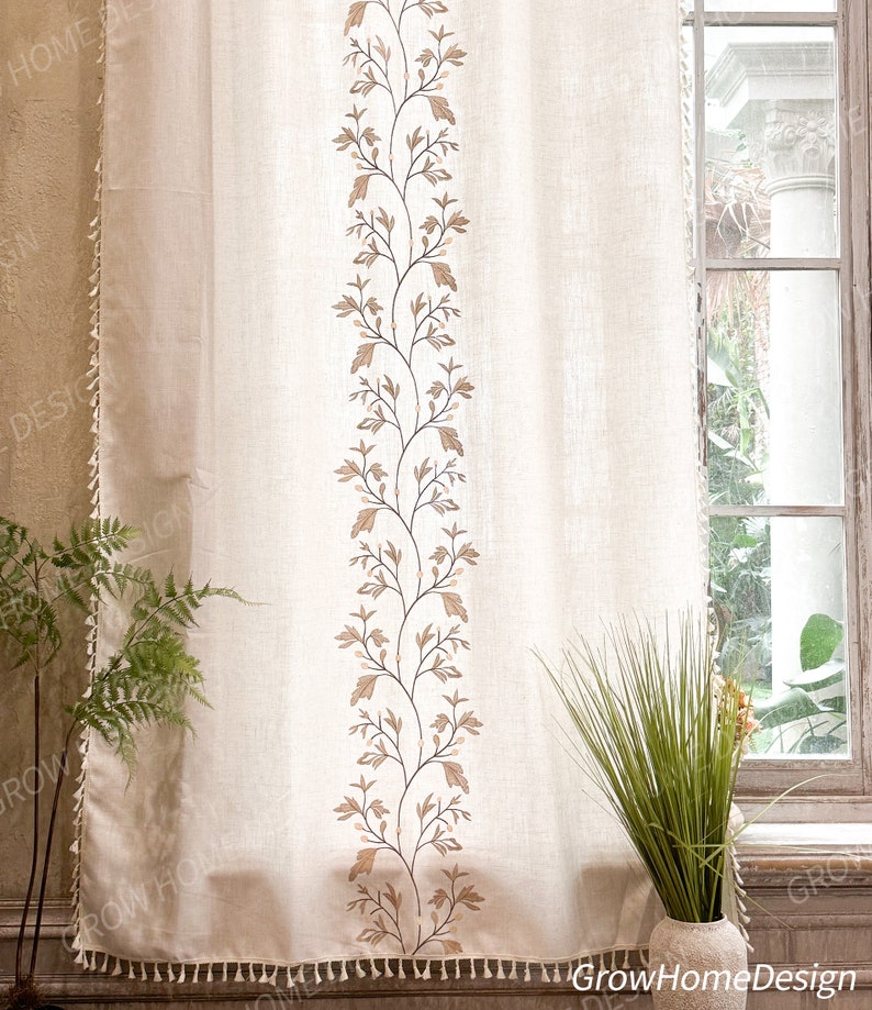 May include: Beige linen curtain with a vertical embroidered floral design in brown and tan. The curtain has a tassel trim along the bottom edge and a matching trim on the left side. The curtain hangs in front of a window.