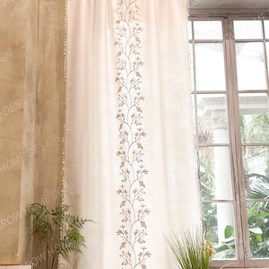 May include: Cream-colored curtain with a central embroidered design of brown leaves and vines. The curtain hangs from a metal rod and has a textured appearance. The design is framed by a decorative edge. The curtain is next to a window.
