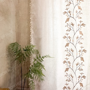 May include: Off-white curtain with a vertical embroidered design of brown branches and leaves. The curtain has a fringe trim on the left edge. A green fern plant is in the lower left corner, and the wall is a light beige color.