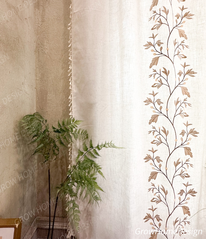 May include: Off-white curtain with a vertical embroidered design of brown branches and leaves. The curtain has a fringe trim on the left edge. A green fern plant is in the lower left corner, and the wall is a light beige color.