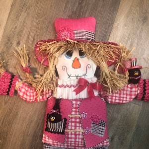 May include: A pink and black checked scarecrow with a pink hat and black birds on its chest. The scarecrow has straw-like hair and a painted face.