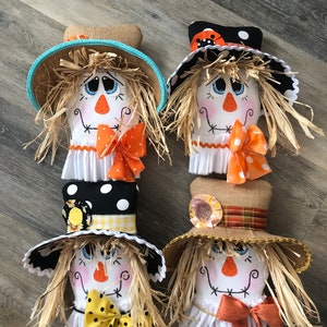 May include: Four handmade scarecrow heads with different hats and bows. The scarecrows have painted faces with black eyes, a red nose, and a stitched mouth. The hats are made of burlap, black and white polka dot fabric, and yellow and white checked fabric. The bows are made of orange, red, and yellow fabric.