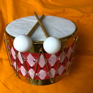 May include: A small red and white drum with gold trim and a diamond pattern. The drum has two white drumsticks with wooden handles.
