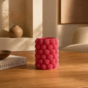 Può includere: Un vaso cilindrico di un vivace colore rosa acceso, caratterizzato da un esterno testurizzato composto da numerose piccole protuberanze sferiche simili a bolle. Ogni sfera presenta un fine motivo a righe concentriche sulla superficie.