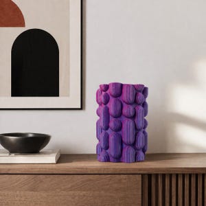 May include: A cylindrical, purple and pink decorative vase with a textured, layered design. The vase is made of vertical, rounded shapes, creating a unique, modern aesthetic. It sits on a wooden surface, with a black bowl and artwork in the background.
