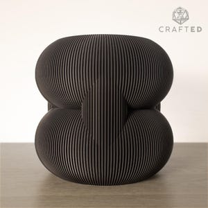 Può includere: Uno sgabello decorativo nero dal design moderno. Presenta una struttura a strati con linee verticali, creando una texture a coste. La forma combina elementi arrotondati, collegati da un elemento centrale. Ideale per interni contemporanei.