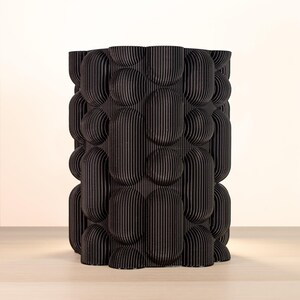 May include: A black, cylindrical decorative vase with a textured, bubble-like pattern. The vase is made of vertical, ridged panels, creating a unique, three-dimensional design. The vase is approximately 10 inches tall.