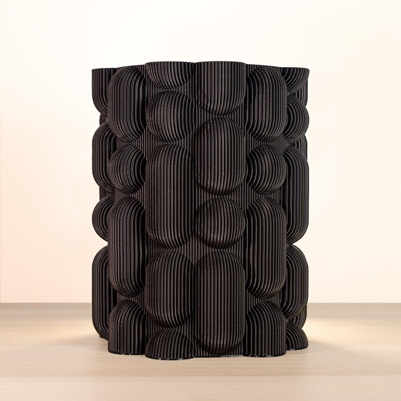 May include: A black, cylindrical decorative vase with a textured, bubble-like pattern. The vase is made of vertical, ridged panels, creating a unique, three-dimensional design. The vase is approximately 10 inches tall.