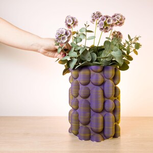 May include: A purple and gold decorative vase with a unique, textured design. The vase is filled with purple flowers and green foliage. The vase is made of layered material and sits on a wooden surface.