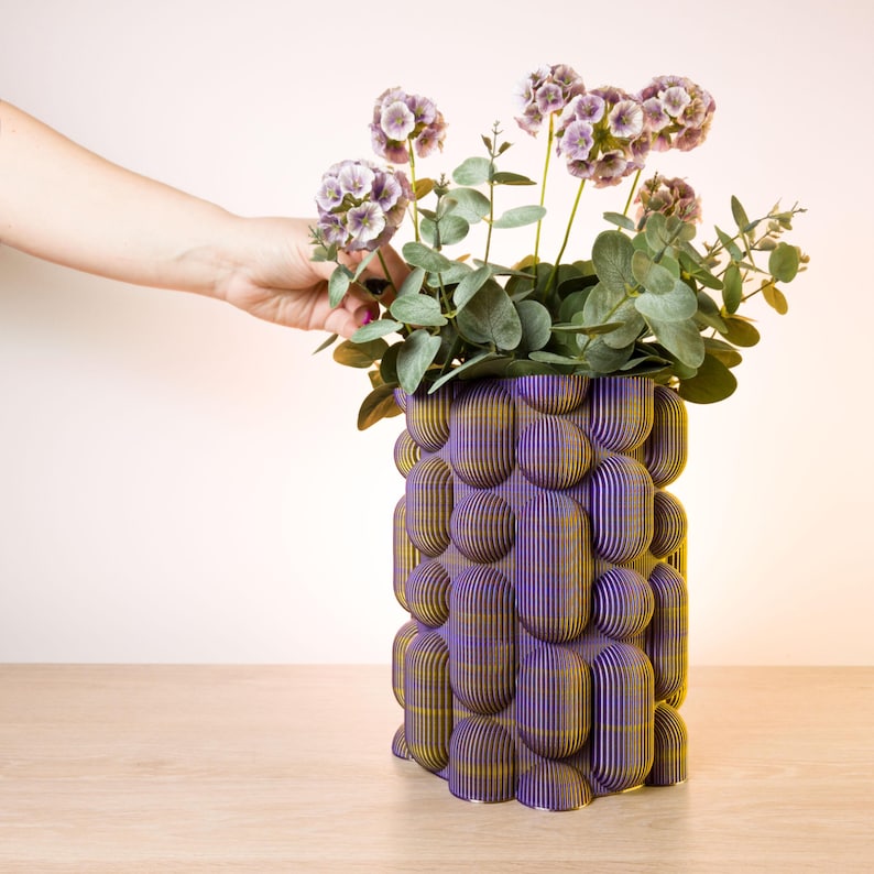 May include: A purple and gold decorative vase with a unique, textured design. The vase is filled with purple flowers and green foliage. The vase is made of layered material and sits on a wooden surface.
