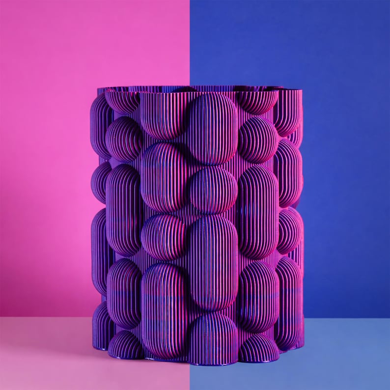 May include: A cylindrical, purple vase with a textured, layered design. The vase features a pattern of rounded, vertical shapes and spheres. The background is split between pink and blue.