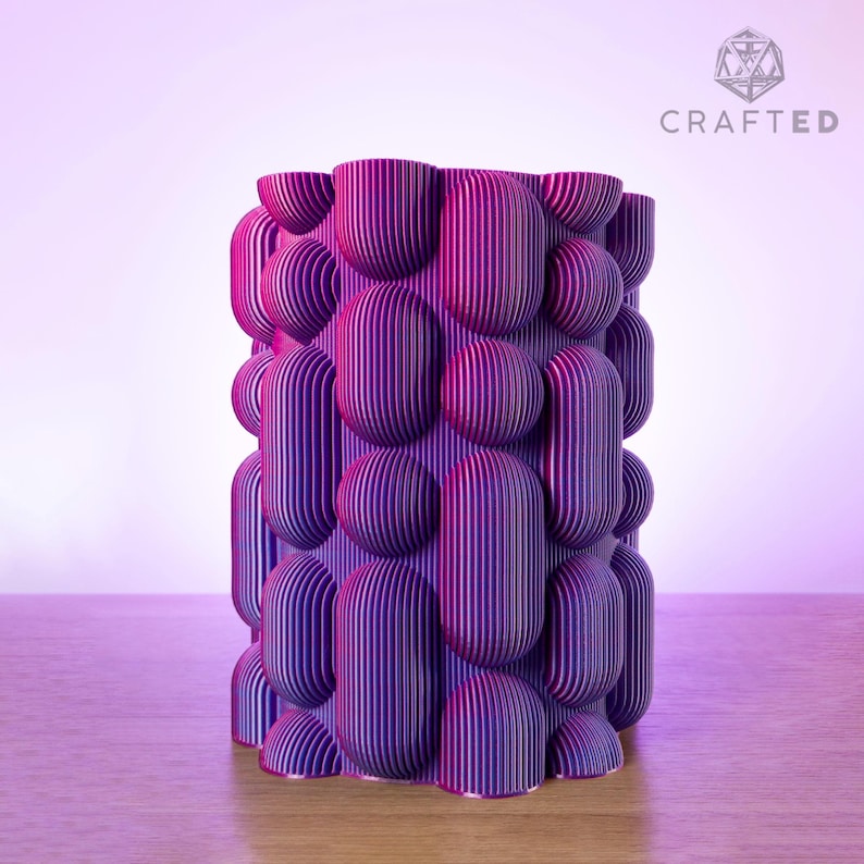 May include: A cylindrical decorative vase with a textured surface. The vase is primarily purple with vertical striped detailing and rounded shapes. The word "CRAFTED" is in the upper right corner.