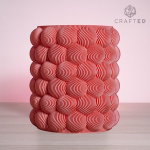 Può includere: Un vaso cilindrico color corallo con un motivo a bolle strutturato. Il vaso presenta una serie di forme arrotondate e stratificate con linee ondulate verticali. La parola "CRAFTED" è stampata in bianco nell'angolo in alto a destra.