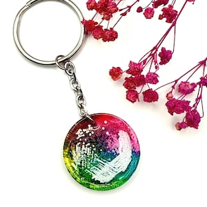 May include: A round keychain with a colorful, abstract design. The keychain features a silver-colored ring and chain. The pendant has a gradient of colors, including red, pink, green, and blue, with white accents. Dried pink flowers are in the background.