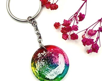 Può includere: Un portachiavi rotondo con un design astratto e colorato. Il portachiavi presenta un anello e una catena color argento. Il ciondolo ha un gradiente di colori, tra cui rosso, rosa, verde e blu, con accenti bianchi. Fiori rosa secchi sullo sfondo.