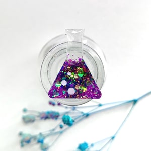 May include: A small, clear glass beaker filled with purple, green, and gold glitter. The beaker is sitting on a white surface.