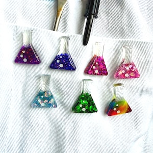 May include: Seven small, clear resin pins shaped like Erlenmeyer flasks, each filled with different coloured glitter and sequins. The colours include purple, blue, magenta, pink, teal, green, and a multi-coloured rainbow blend, all featuring white circular accents.