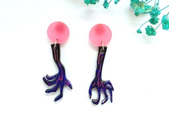 Può includere: Un paio di orecchini unici con un perno circolare rosa, uno stelo nero e viola e un design a forma di mano. Gli orecchini sono realizzati in un materiale lucido e sono su uno sfondo bianco.