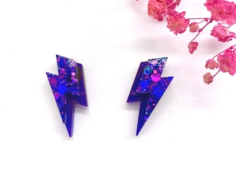 May include: A pair of lightning bolt shaped earrings in a deep purple hue. The surface of each earring is embedded with iridescent blue and purple glitter particles of varying sizes, creating a reflective and textured appearance on the front.