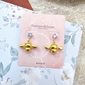 May include: Gold-tone earrings featuring a small cartoon dog charm with a clear rhinestone dangling from the top. The earrings are displayed on a pink card with the text ".thetrendblazer. curated collection #loveyourself"