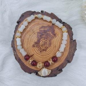 May include: A white and red beaded bracelet with a gold charm in the shape of a fan. The bracelet is on a wooden surface.