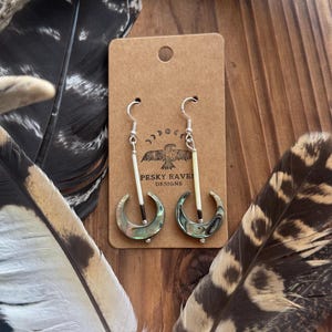 Può includere: Orecchini in argento con un design a mezzaluna, con conchiglie di abalone iridescenti. Ogni orecchino ha un accento cilindrico bianco e una piccola perla d'argento. Gli orecchini pendono da ganci d'argento. Il testo "PESKY RAVEN DESIGNS" è visibile.