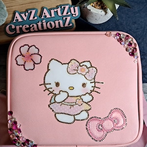 May include: Pink Hello Kitty laptop case with a pink flower, a pink bow, and rhinestones. The case has the text "AvZ Artzy CreationZ" on the front.