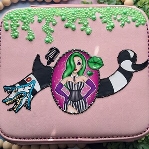 May include: Pink rectangular box with a cartoon design featuring a woman with green hair, a black and white striped outfit, and a shark-like creature. The box is decorated with green pearl-like accents and a black and white striped design.