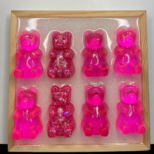 Op de afbeelding: Een houten frame met negen roze gummy bear sculpturen, sommige met glitter, gerangschikt in een rasterpatroon op een witte achtergrond.