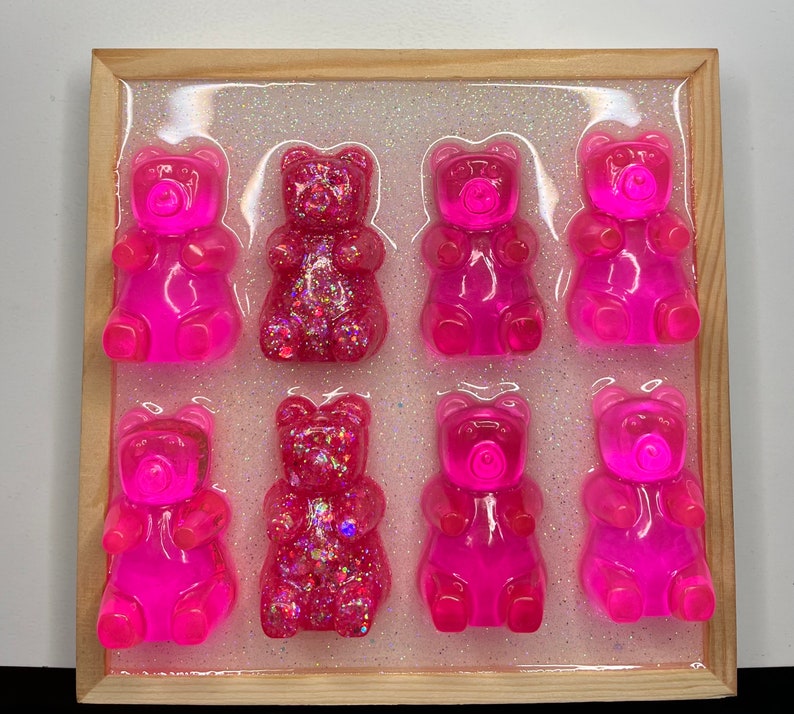 Op de afbeelding: Een houten frame met negen roze gummy bear sculpturen, sommige met glitter, gerangschikt in een rasterpatroon op een witte achtergrond.