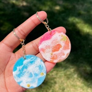 May include: Two round resin keychains with a swirling white and blue design and a swirling white and pink design. Both keychains have gold glitter embedded in the resin.