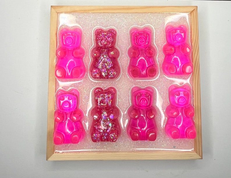 Op de afbeelding: Een houten frame met een transparante achtergrond en acht roze gummybeertjes. De gummybeertjes zijn gerangschikt in twee rijen van vier. Sommige van de gummybeertjes hebben glitter erin verwerkt.