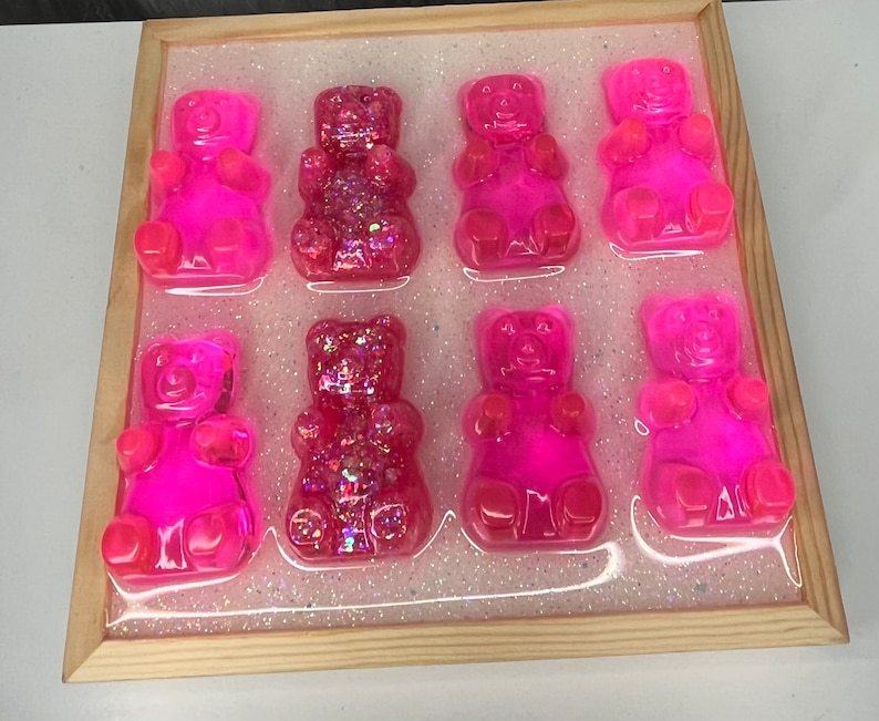Op de afbeelding: Negen roze gummybeertjes in de vorm van beren met glitter erin verwerkt. De gummybeertjes zijn gerangschikt in een raster op een witte achtergrond met glitter.