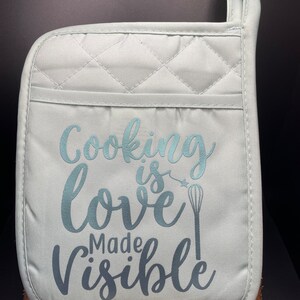 May include: A light blue quilted oven mitt with the text "Cooking is love made visible" and a whisk graphic.