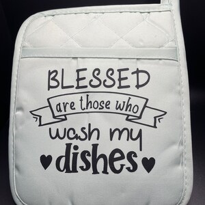 May include: A light green quilted pot holder with a black design that reads "Blessed are those who wash my dishes" with a heart on each side of the word "dishes".