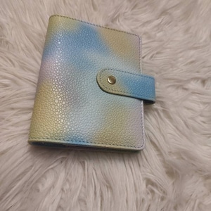 Può includere: Un piccolo taccuino con motivo tie-dye pastello e chiusura a bottone dorato.