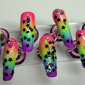 Può includere: Unghie press-on con sfumatura arcobaleno e glitter nero a forma di stella. Le unghie sono lunghe e a forma di bara.