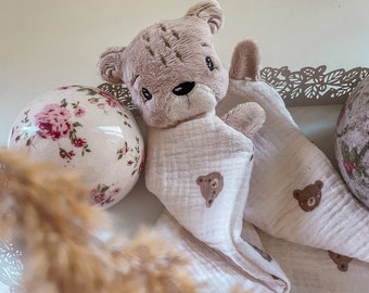 May include: A plush brown teddy bear wrapped in a white muslin swaddle blanket with a brown bear print. The blanket is decorated with small brown bears. The teddy bear is lying on a white surface with a floral patterned ball and a white lace tray.