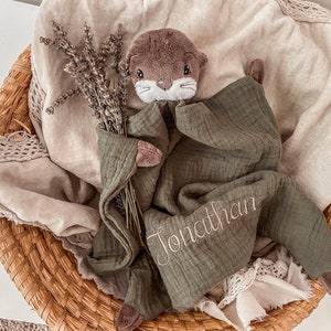 May include: A green muslin baby blanket with a brown plush otter toy. The blanket has the name "Jonathan" embroidered on it.