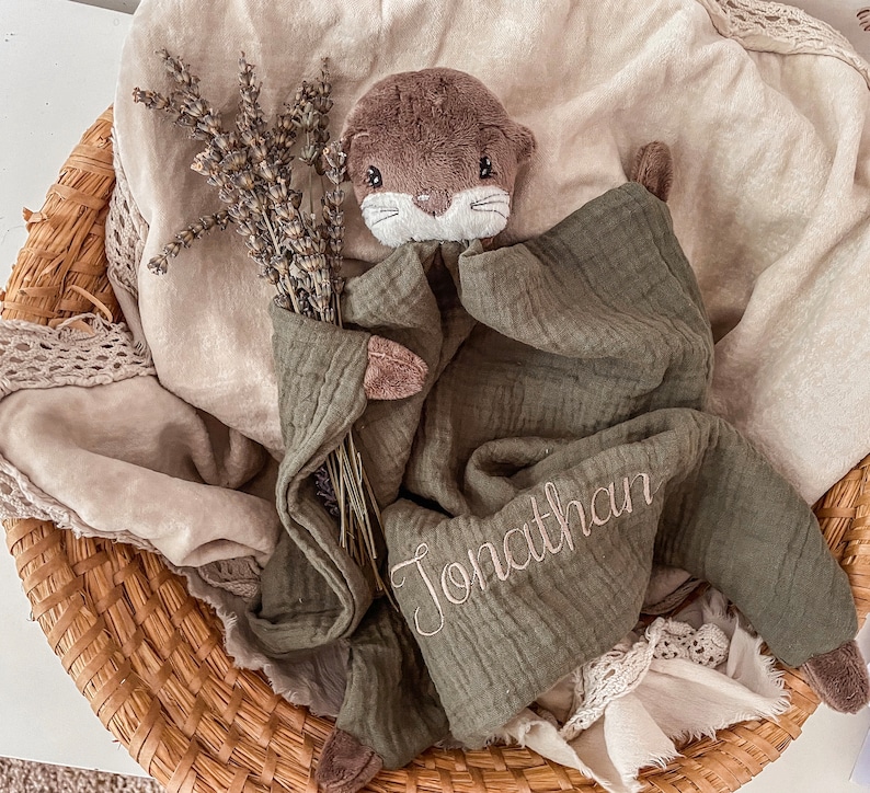 May include: A green muslin baby blanket with a brown plush otter toy. The blanket has the name "Jonathan" embroidered on it.