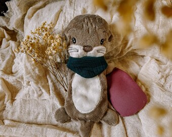 May include: A plush otter toy with a gray body, white belly, and a dark teal scarf. The otter has embroidered facial features and is accompanied by a burgundy-colored pillow and a sprig of dried flowers. The toy is on a cream-colored fabric.