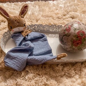 May include: A blue muslin baby blanket with a brown bunny lovey on a white metal tray with a floral patterned ball.
