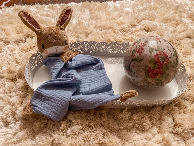 May include: A blue muslin baby blanket with a brown bunny lovey on a white metal tray with a floral patterned ball.