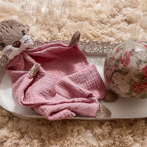 May include: A pink muslin baby blanket with a brown plush otter head attached. The blanket is lying on a white tray with a floral patterned ball beside it.