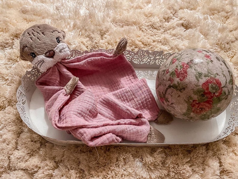 May include: A pink muslin baby blanket with a brown plush otter head attached. The blanket is lying on a white tray with a floral patterned ball beside it.
