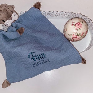 May include: A blue muslin baby blanket with a brown otter stuffed animal attached. The blanket has the name "Finn" and the date "20.01.2023" embroidered on it.