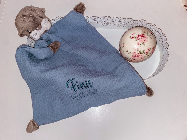 May include: A blue muslin baby blanket with a brown otter stuffed animal attached. The blanket has the name "Finn" and the date "20.01.2023" embroidered on it.