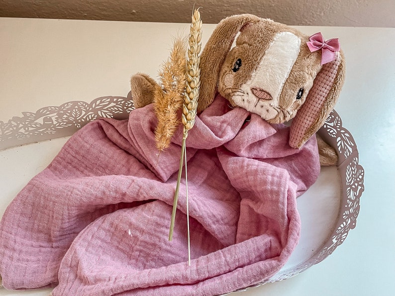 May include: A plush brown and white bunny with a pink bow sits on a pink muslin blanket in a decorative metal tray. A dried wheat stalk is placed next to the bunny.