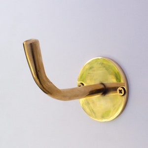 May include: A brass wall hook with a rounded base and a single hook extending out. The hook is mounted on a white wall.