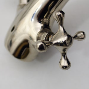 Pode incluir: Close-up de uma pega de torneira cromada com um design em forma de cruz. A pega &eacute; brilhante e reflete a luz.